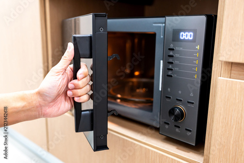 Hand holding door handle opening black microwave oven in modern wooden cabinet. Home appliance in the kitchen