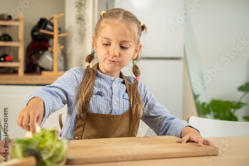 Cute little girl pressing a bread slice on wooden cutting board while making sandwich in kitchen. Adorable caucasian child enjoying preparing healthy breakfast snack at home kitchen table.