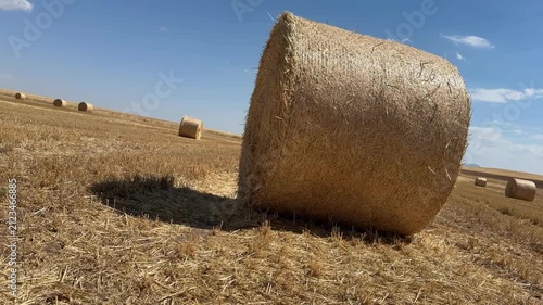A cinematic 4K low-angle tracking shot of a large round hay bale in a golden harvested wheat field under a bright blue summer sky with scattered white clouds.