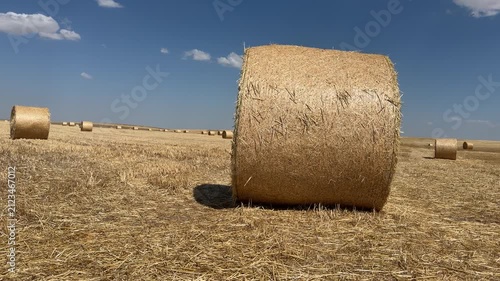 A cinematic 4K low-angle tracking shot of a large round hay bale in a golden harvested wheat field under a bright blue summer sky with scattered white clouds.