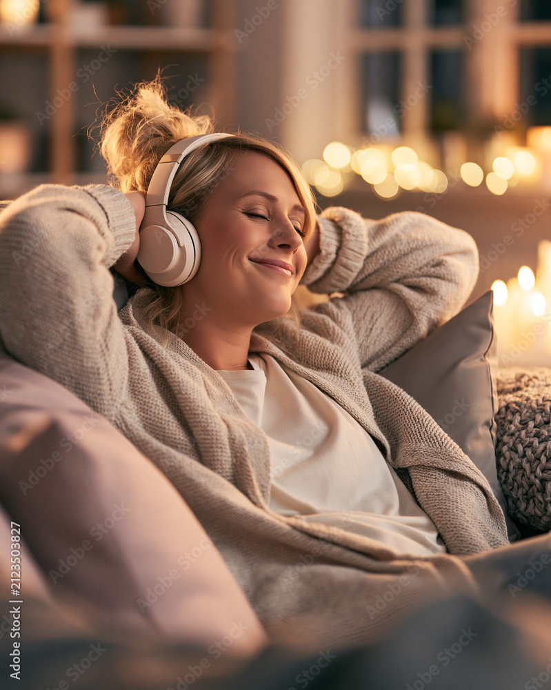 Obraz premium A relaxed woman with blonde hair in a messy bun wears wireless headphones leaning back on a sofa while enjoying music in a cozy room