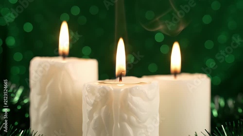 Three White Candles Lit With Gentle Flames And Wisps Of Smoke Against A Blurry Green Background With Bokeh And Festive Tinsel