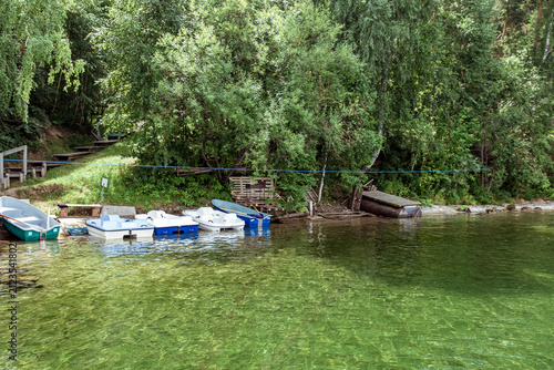 A row of colorful catamarans, pedal boats, and small rowboats, docked at a rustic wooden pier on a clear green lake surrounded by a dense birch forest and a grassy slope