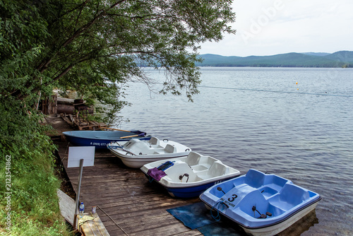 Water bikes and catamarans lined up on a wooden dock by a peaceful lake with lush greenery and distant hills are perfect for relaxation.