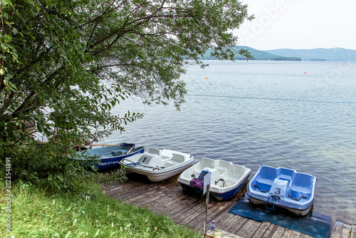 Water bikes and catamarans lined up on a wooden dock by a peaceful lake with lush greenery and distant hills are perfect for relaxation.