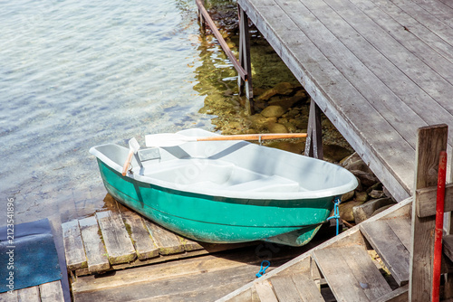 Turquoise and white plastic rowboat resting on wooden planks beside a weathered dock, clear shallow water reveals smooth river stones, wooden oar rests across the boat, rustic lakeside scene