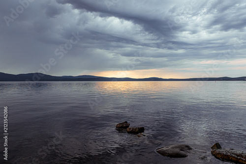 Dramatic cloudy sky over a calm lake with silhouetted hills, warm sunset glow reflecting on water, and wet rocks in the foreground, capturing moody natural beauty.