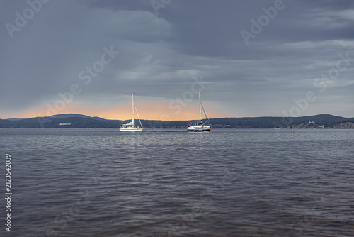 Sailboats anchored on Lake under a moody overcast sky, with distant hills and a subtle sunset glow on the horizon, conveying a tranquil yet dramatic atmosphere