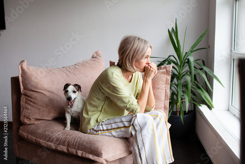 Caucasian woman sitting on couch looking out window with dog. Sad girl feeling lonely at home. Concept of depression or melancholy and support from loyal pet. Emotional domestic scene.
