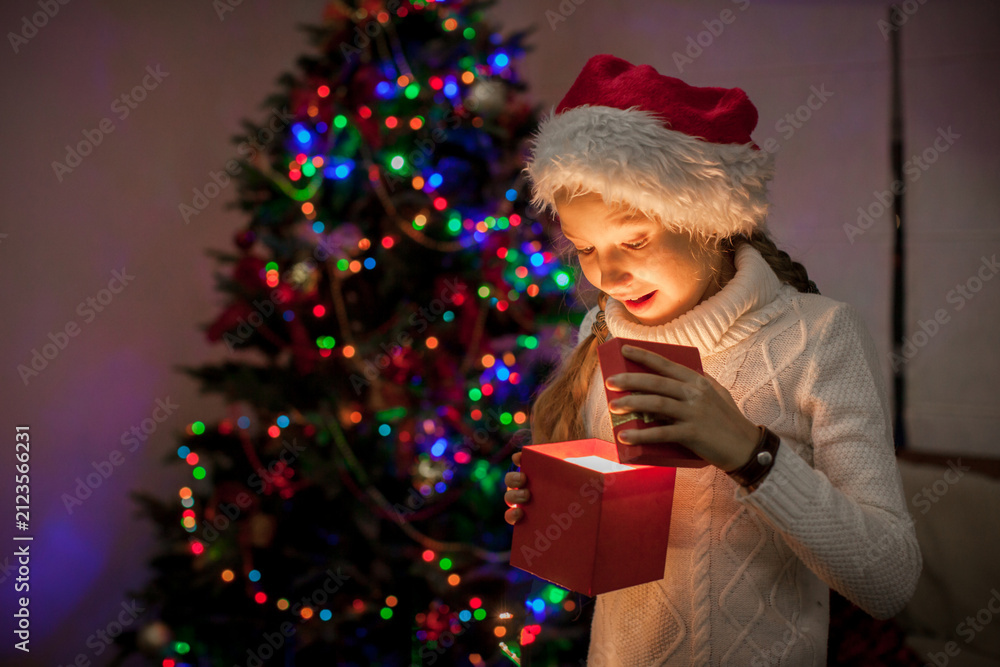 Obraz premium Child opening glowing gift box near decorated Christmas tree. Happy girl wearing Santa hat receiving present. Magic of holiday season and winter celebration at home with light reflection.