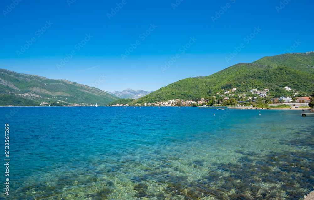 Naklejka premium Clear blue sea meets green hillside under bright blue sky in Tivat, Montenegro, with shoreline featuring small houses and rocky beach.