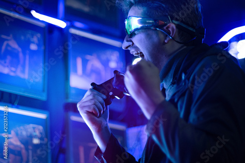 Male gamer, wearing headphones, is playing online video games or competing in esports, displaying a triumphant gesture in a neon-lit gaming room.
