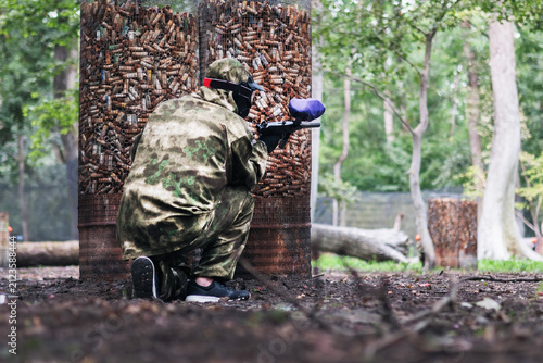 Paintball player with marker standing among old barrels in the forest