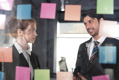 Manager makes a skeptical face at a sticky note while the young woman in profile keeps scanning the colorful plan on the glass wall in a bright modern coworking office.