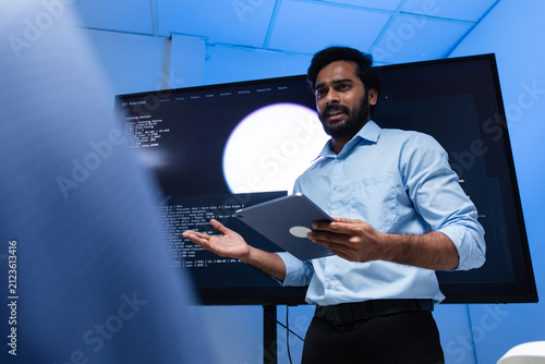 Professional Indian Male Software Engineer Presenting Code and AI Data to Team in Modern Tech Office, Indian Male Programmer Collaborating with Development Team and Reviewing Code on Computer Screen.