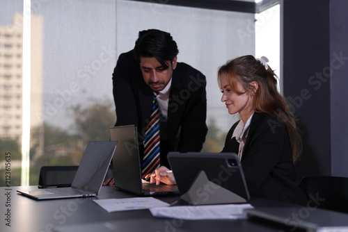 Konstfotografi Smiling seated businesswoman with long strawberry blonde hair follows her manager's fingertip as he indicates a line on the laptop beside her tablet stand in a bright modern coworking