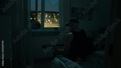 
A man sits in a room in the evening darkness, playing the guitar, with a view of the traffic outside.
The slider shot slowly moves closer.