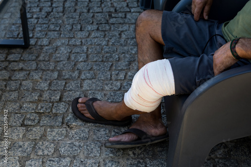 Side view of a man sitting outdoors on a chair, using his mobile phone while resting his bandaged knee joint on cobblestone pavement.