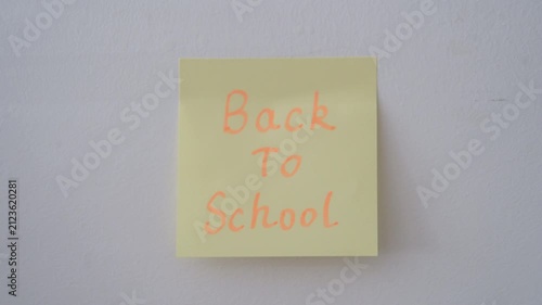 A person's hand pressing a square yellow sticky note onto a plain white wall. The note features the words 