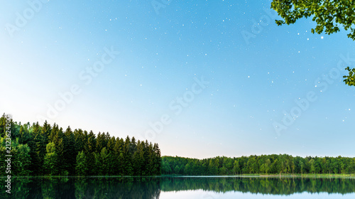 Serene landscape featuring calm lake reflecting clear sky filled with stars, surrounded by lush green trees. tranquil atmosphere evokes sense
