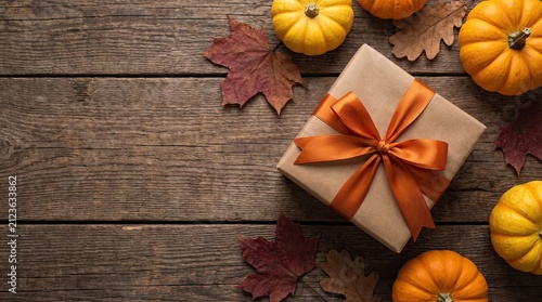 A wrapped gift box decorated with an orange ribbon surrounded by small pumpkins and autumn leaves on rustic wooden background