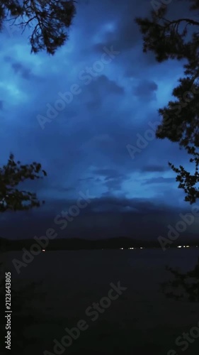 Lightning flashes in the storm clouds, illuminating the mountain lake. In the foreground, trees sway in the wind. Blue twilight sky, thunderstorm, twilight