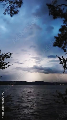 Lightning flashes in the storm clouds, illuminating the mountain lake. In the foreground, trees sway in the wind. Blue twilight sky, thunderstorm, twilight