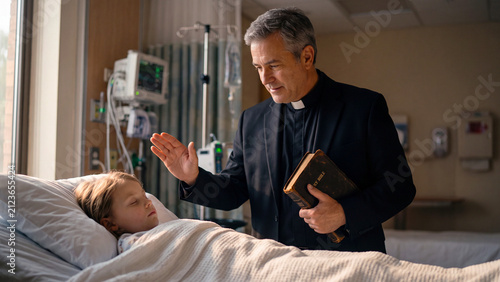 Priest holding a bible and blessing a young patient lying in a hospital bed, faith and healing