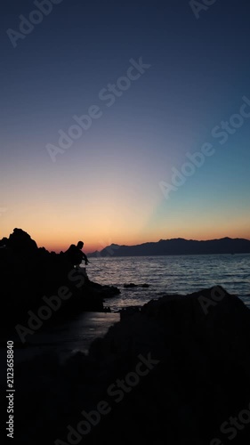 Vertical screen Spinning fishing at sunset follows an angler retrieving lure across calm water as fading daylight creates peaceful evening atmosphere by rocky shore