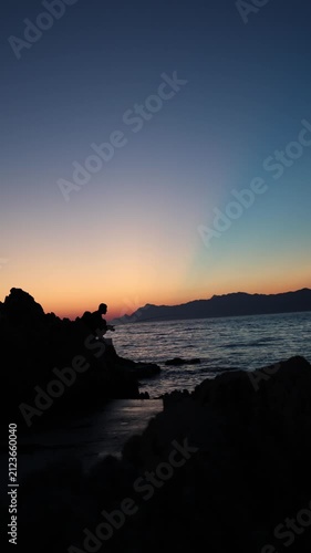 Vertical screen Sunset fishing silhouette on rugged rocks creates peaceful evening mood with calm waves fading light and quiet reflection after a long summer day