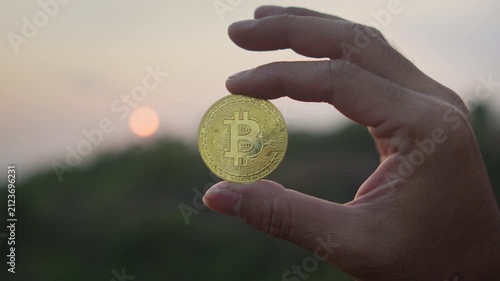 Close up of a hand holding a physical Bitcoin coin against a dramatic sunset sky, symbolizing digital finance, investment, and the future of decentralized currency.