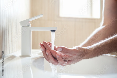 Close up shot of man washing hands in sink under running tap water with modern white faucet in bright bathroom environment