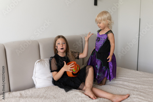Girls in witch costumes gesturing surprised on bed with pumpkin bucket