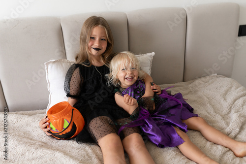 Two sisters in witch costumes hugging and laughing on bed with pumpkin bucket