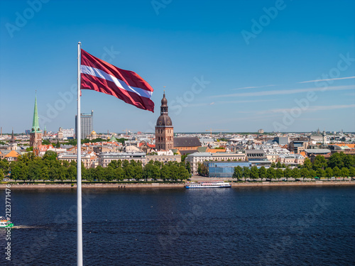 Konstfotografi Aerial view of the red-white-red Latvian flag waving in the wind high above the Daugava, framing the UNESCO-listed Old Town of Riga, under a clear blue summer sky