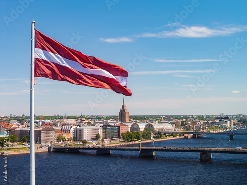 Foto Aerial drone view of the Latvian flag waving over the wide Daugava, backed by the bridges and skyline of central Riga, under a clear blue summer sky