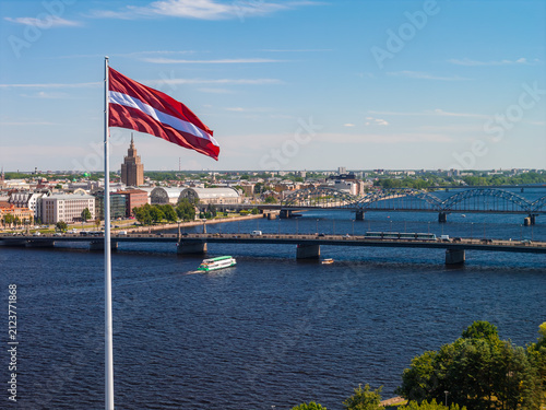 Foto Aerial drone view of the Latvian flag waving over the wide Daugava, backed by the bridges and skyline of central Riga, under a clear blue summer sky