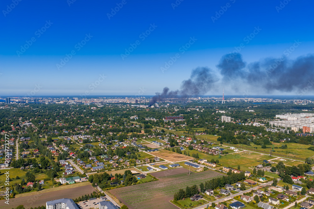 Fototapeta premium Aerial view of a thick column of black smoke from a large fire rising high above the rooftops and green districts of Riga, in clear daylight.