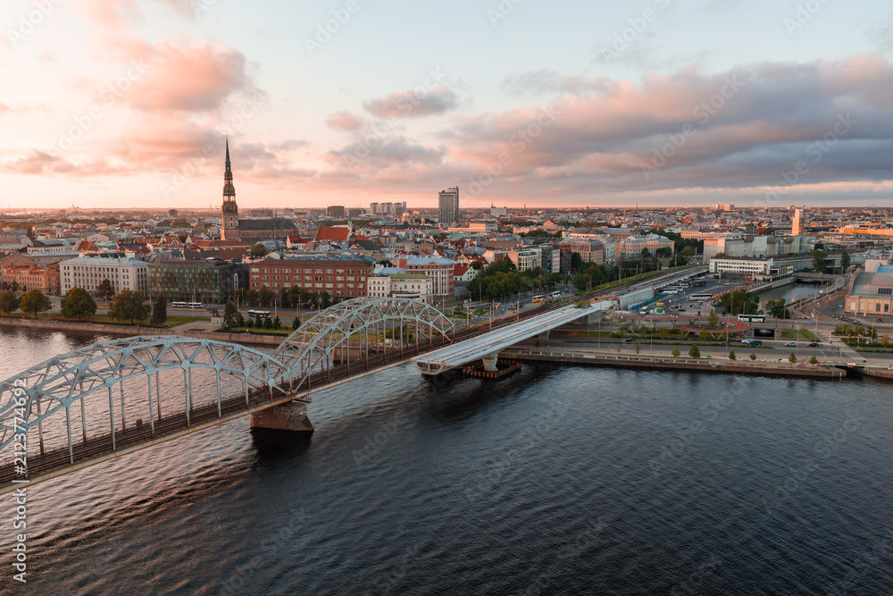Obraz premium Aerial drone view of the riveted steel arches of the Riga railway bridge spanning the wide Daugava river towards the city centre, at sunset.