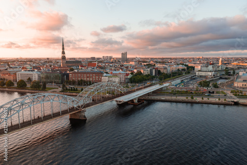 Aerial drone view of the riveted steel arches of the Riga railway bridge spanning the wide Daugava river towards the city centre, at sunset.