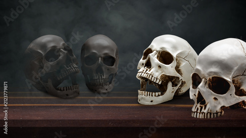 Konstfotografi Four human skulls, two black and two white, arranged on a dark wooden table against a moody, textured background, representing themes of life, death, duality, and mortality