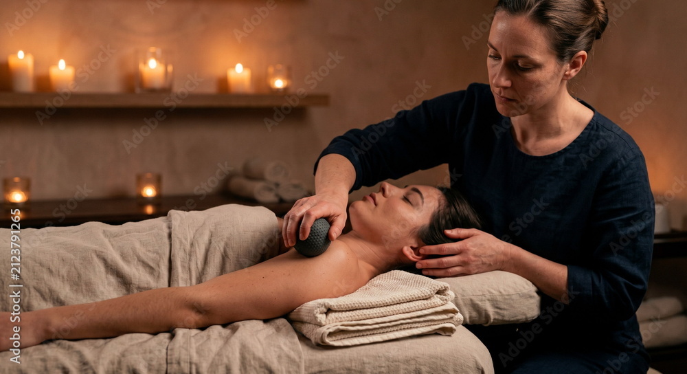 Fototapeta premium Manual therapist performs myofascial release on a client shoulder with a massage ball and towels in a serene spa treatment room.