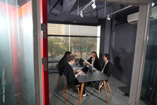 Foto A tinted office partition frames four coworkers deep in discussion at a laptop table, warm daylight glowing through the tall coworking window, reviewing plans in warm natural daylight