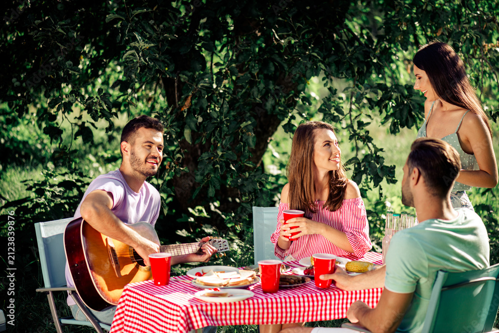 Fototapeta premium Friends enjoying a summer picnic outdoors with guitar, food and red cups at a checkered tablecloth under trees