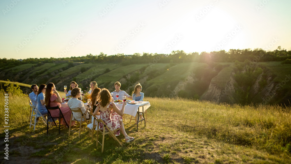 Obraz premium Friends gathered around outdoor dinner table in vast sunlit Riviera landscape. Concept of scenic lifestyle, resort event planning, nature-based hospitality.