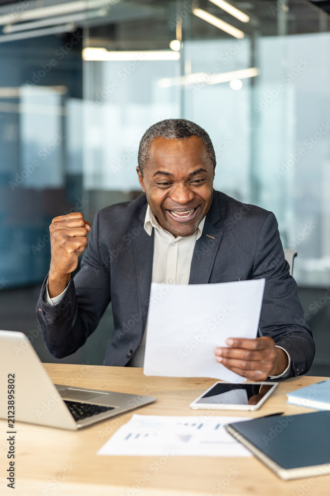 Obraz premium African american businessman celebrating success and achievement, reading a document with an excited expression and clenched fist while sitting at a modern office desk with a laptop and tablet