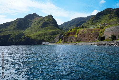 Bild auf Leinwand Beautiful Madeira coastline with dramatic green mountains, volcanic cliffs, a coastal road and the deep blue Atlantic Ocean under a bright blue sky