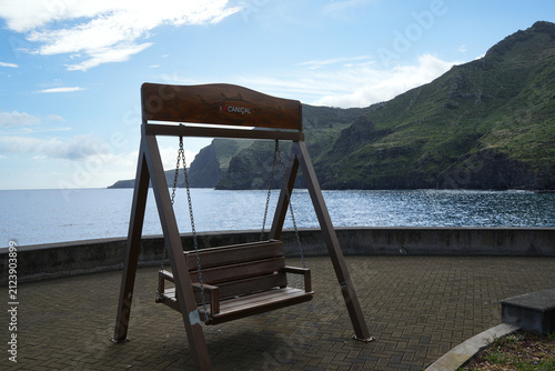 Foto Wooden swing overlooking the Atlantic Ocean and dramatic green mountains in Caniçal, Madeira, Portugal, with a peaceful coastal setting and beautiful island scenery