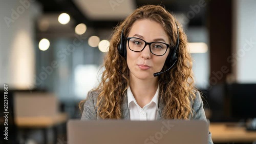 Woman with curly hair wearing headset and glasses talking while looking at camera. Professional office setting with laptop. Concept of customer service, call center, remote work, technical support.