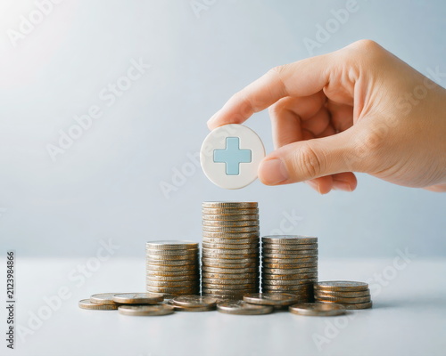 Health insurance, hand placing medical cross symbol on increasing coin stacks, light background, financial planning for healthcare.